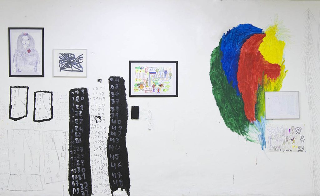 Цветная горизонтально-ориентированная фотография. Белая стена выставочного пространства. На ней росписи художников Артема Левинского — яркая разноцветная абстракция, и Сергея Чубарова — черно-белые цифры. Хаотично висит графика в рамах.